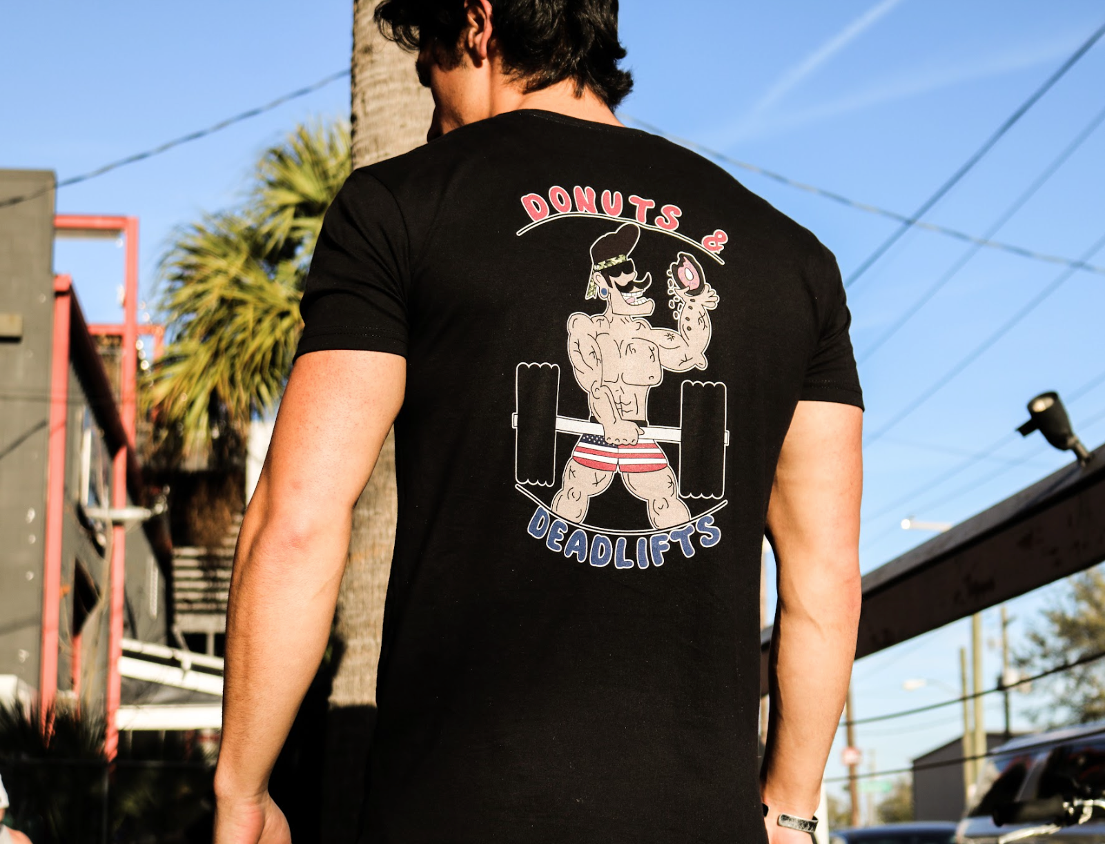 Man wearing black T-Shirt with the "Donuts & Deadlifts" written on the back & a character figure eating a donut while doing a deadlift